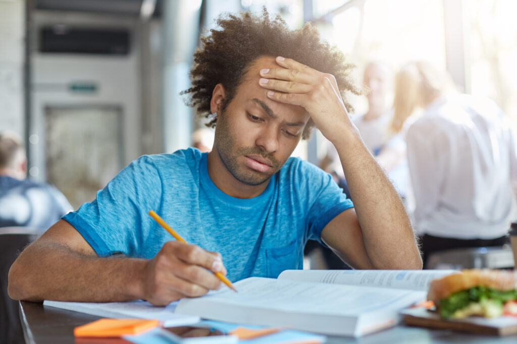 Exhausted male student with bristle, dark skin and fluffy hair sitting at university canteen eatting hamburger preparing for exams holding hand on forehead trying to concentrate on his writing