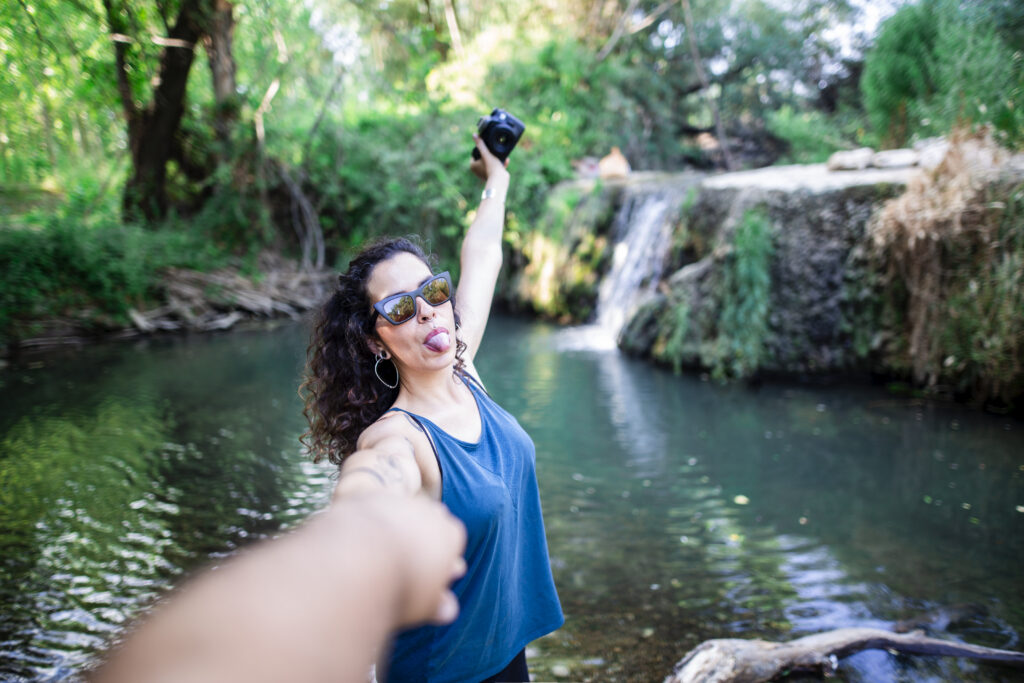 Girl showing her tongue holding her partner's hand and taking a selfie of herself at a waterfall