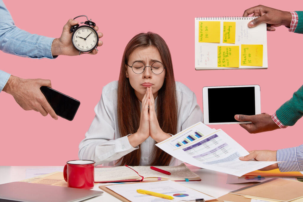 Wishful college student keeps hands in praying gesture, believes in good fortune, closes eyes, prepares task in economics, asks God dreams come true, isolated over pink background, works overtime